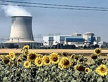 Photo avec tournesols à l'avant et une usine sur le dos. L'usine a une cheminée fumante large avec un diamètre comparable à sa hauteur.