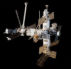 Une vue de Mir en toile de fond la branche de la Terre. Compte tenu quatre modules cylindriques couverts dans l'isolation blanches disposées en forme de croix sur une petite sphère centrale. Un autre des projets du module arrière de ce domaine, et un petit module est fixé à l'extrémité de cette. Chaque module est la germination différents panneaux solaires, des grues et autres équipements grêles, avec Soyouz et Progress vaisseau amarré aux ports du complexe avant et arrière.