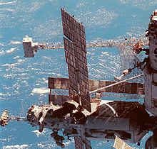 Une vue partielle d'une station spatiale, avec deux modules visibles. Le module aligné verticalement a une grande antenne, en forme d'assiette en saillie de celle-ci, et un grand treillis peut être vu à l'horizontale derrière elle, avec un dispositif en forme de boîte blanc monté sur l'extrémité. Le module comporte un treillis horizontal inférieur monté à l'extrémité de celui-ci, avec un sac à dos blanc jointe à la présente. Le module comporte également deux grands panneaux solaires et un certain nombre de caméras montées sur une unité sur l'extrémité distale de celui-ci. Un autre panneau solaire est visible derrière ce module.