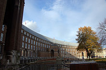 Un grand bâtiment en briques, construit dans une courbe peu profonde, avec un porche central. Devant qu'une piscine et une fontaine d'eau. arbres d'automne sur le droit et un ciel bleu avec quelques nuages au-dessus.