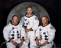 Trois astronautes en scaphandres sans casques, assis devant une grande photo de la lune.