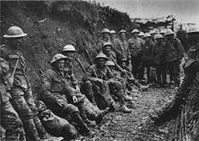 Photo en noir et blanc de deux douzaine d'hommes en uniformes militaires et des casques métalliques assis ou debout dans une tranchée boueuse.