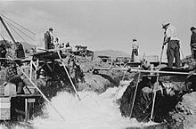 Quatre hommes vêtus de chemises à manches longues, un pantalon long, et les chapeaux sont perchés sur les plates-formes des deux côtés d'un torrent. Trois des hommes sont debout, et l'on est assis. Chaque homme tient une extrémité d'une longue perche avec un filet, trempé dans l'eau, attaché à l'autre extrémité. Plusieurs personnes sans pôles regardent ou en attente à proximité.