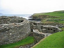 Un mur semi-circulaire pierre conseils gauche à l'existence d'un grand bâtiment ancien et à droite se trouvent les ruines de diverses autres structures de pierre. Au fond, un falaise basse divise un corps d'eau de champs herbeux.