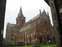 Une grande église en pierre rouge et jaune avec une tour carrée et une flèche sur la tour.