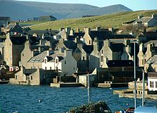 Les maisons de pierre se pressent autour d'un rivage, le pignon extrémités face à l'eau, avec des collines vertes au-delà.