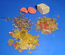 Sur un fond bleu, deux grandes plaquettes rouges légers en haut à gauche, un cube comprimé gris clair en haut à droite, les petites pastilles brunes au milieu à gauche, et, au milieu et en bas à droite, divers flocons jaune-vert et rouge