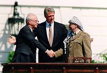 Un homme en costume sombre sur la gauche serre la main d'un homme portant une coiffure traditionnelle arabe sur la droite. Un autre homme (Bill Clinton) se tient avec les bras ouverts dans le centre derrière eux.
