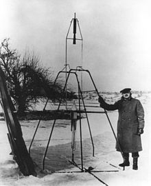 Structure de châssis en métal situé sur la neige près d'un arbre. Un homme d'âge moyen portant un manteau, des bottes, des gants de cuir et un chapeau se en tient à la structure et le tient avec sa main droite.