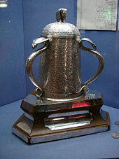 Dans une vitrine bleu est une forme de cloche coupe d'argent qui est fortement gravé avec des motifs; elle est ornée sur son couvercle avec un petit éléphant d'argent et deux de ses grandes poignées sont cobras argent.