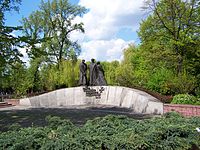 Un muret de pierre, se courbant vers le haut. Three statues of Polish soldiers are mounted at its center. Below the statues, Text is mounted as per caption.