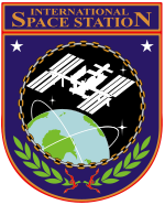 Une silhouette de l'ISS en orbite montré dessus de la Terre. Cette image est suspendu dans un bouclier orange et violet, avec de la Station spatiale internationale »les mots ci-dessus l'image, et de feuilles de laurier-dessous.