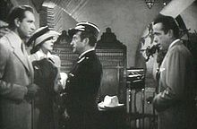Noir et blanc film de capture d'écran de plusieurs personnes dans une discothèque. Un homme à l'extrême gauche est vêtu d'un costume et a une femme debout à côté de lui porter un chapeau et robe. Un homme au centre est à la recherche à l'homme sur la gauche. Un homme à l'extrême droite est vêtu d'un costume et qui cherchent à les autres personnes.