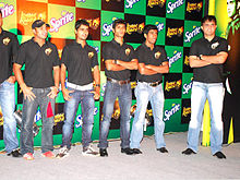 Un groupe d'hommes debout, portant des T-shirts et bleu noir jean délavé. Chacun d'entre eux se tournent vers la gauche de l'image. The backdrop has alternate black and green boxes.