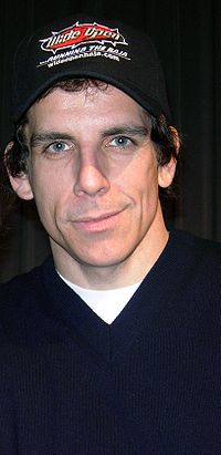 Stiller est face à la caméra et souriant. Il porte une casquette de baseball et une chemise bleu avec un t-shirt blanc en dessous.