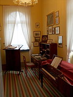 chambre meublée dans le style début du XIXe siècle, avec des rayures coureur sur le plancher; murs peints de pêche et le mur à droite ornés de petits tableaux; dans la droite au premier plan un piano carré, à l'arrière gauche devant une fenêtre d'un stand de lecture, à l'arrière droit d'un bureau, tout en bois sombre