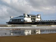Une passerelle soutenue par des pieds métalliques provenant de la mer, à l'extrémité duquel se trouve un grand bâtiment blanc avec un toit et coin tourelles en forme de vague.