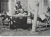 Trois hommes sont assis sur des chaises devant une maison, un homme chauve dans la quarantaine est assis dans le centre, à côté d'une table entre les deux jeunes hommes. L'homme sur le droit a ses pieds sur la table et tient un scénario. le vieil homme est d'avoir essuyé son front par une infirmière qui se tient sur lui.