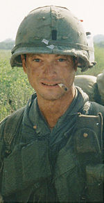 Un homme est au centre de l'image en regardant la caméra. Il est vêtu Vietnam-ère tenue militaire, y compris un gilet et casque. Il a une cigarette assis sur ses lèvres et porte un sac à dos.