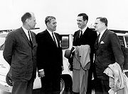 Quatre hommes en costumes sont à l'extérieur, parler à l'autre en face d'un grand automobile blanc.