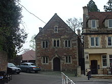 Bâtiment en pierre avec porte voûtée au rez de chaussée. Trois fenêtres au premier étage et la fenêtre unique en dessous du niveau du toit. Pour la gauche sont les arbres et les voitures, à la droite un autre bâtiment.