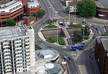 Vue aérienne du rond-point, une jonction de plusieurs rues. Véhicules traversent autour du rond-point, qui est entouré de bâtiments, principalement multi-étages