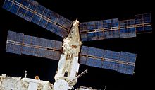 Une vue du module Spektr avec la noirceur de l'espace derrière. Compte tenu quatre panneaux solaires sont dorées du module, en plus du module cylindrique elle-même, recouverte d'un isolant et blanc avec un cône à l'extrémité distale du module. Deux des tableaux de projeter ce cône, et deux projet depuis l'extrémité distale du cylindre.