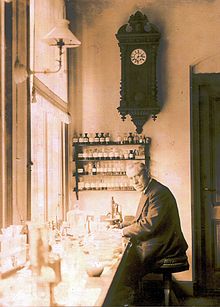 Un vieux, homme à lunettes vêtu d'un costume et assis sur un banc par une grande fenêtre. Le banc est couvert de petits flacons et des tubes à essai. Sur le mur derrière lui, ce est une grande horloge ancienne dessous de laquelle sont quatre étagères fermées petits sur lequel siègent de nombreuses bouteilles soigneusement étiquetés.