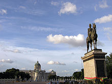 Une grande place avec une statue en bronze d'un homme sur un cheval dans le centre; au-delà de la place est un grand bâtiment de deux étages avec un toit en forme de dôme, fenêtres cintrées et des colonnes
