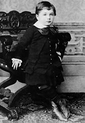 Un jeune garçon aux cheveux courts et un visage rond, portant un collier blanc et grand arc, avec le gilet, manteau, jupe et des bottes hautes. Il est appuyé contre une chaise ornée.