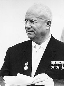 Un coup de portrait d'un homme chauve plus âgé avec des lunettes à double foyer. Il est vêtu d'un blazer sur une chemise à col et une cravate. Dans ses mains, il tient une série de documents.