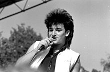 Une image en noir et blanc d'un homme à la peau claire avec un microphone tenu à sa bouche. Il est visible à partir de la poitrine et porte une chemise noire sans manches avec un gilet sans manches blanc ouvert overtop. Une petite croix est portée autour du cou. Ses cheveux noirs sont coiffés en un mulet. L'homme regarde devant la caméra vers la gauche. Un mélange d'arbres et le ciel sont visibles en arrière-plan.