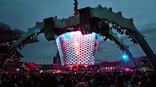 Une scène de concert; quatre grandes jambes courbe au-dessus de la scène et maintenez un écran vidéo qui se étend jusqu'à la bande. Les jambes sont allumés en vert. L'écran vidéo a des lumières multicolores clignotantes sur elle. Le public entoure la scène de tous les côtés.