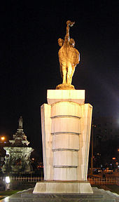Une statue de pierre de porte-flambeaux comme on le voit dans la nuit. Une fontaine avec une base blanche est en arrière-plan