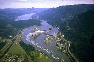 Vue aérienne d'une grande rivière qui serpente à travers une gorge montagneuse. Il passe au-dessus d'un barrage se étend dans quatre segments d'une banque à travers trois îles intermédiaires. Autoroutes, en passant par des grappes de bâtiments ici et là sur les deux rives, sont parallèles à la rivière. Whitewater et de mousse enroulent en aval de l'un des segments centraux.