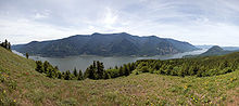 A courbes des rivières larges doucement à la base d'une chaîne de montagne. Une prairie au premier plan cède la place à une forêt de conifères, puis à la rivière. Dans le fond, une couche de nuages minces voile un ciel bleu.