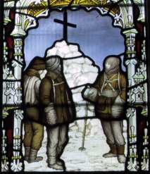 Trois figures sont représentées en verre coloré, debout par un cairn de neige surmonté d'une grande croix. La scène est encadrée par un arc décoratif.