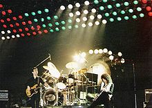 Trois membres du groupe lors d'un concert à Hanovre. De gauche à droite, John Deacon (résisté désinvolture), Roger Taylor (jeu, assis au kit de batterie), et Brian May (semble jouer intensément). Derrière Taylor est un tam-tam utilisé à la fin de Bohemian Rhapsody. Derrière ce est un grand jeu de lumières multicolores soulevées dessus de la scène.