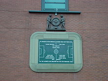 Une tablette de pierre, inscrit avec l'image d'un terrain de football et plusieurs noms. Il est entouré par une bordure en pierre en forme d'un stade de football. Au-dessus de la tablette est une sculpture en bois de deux hommes tenant un grand couronne.