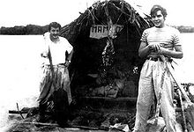 photographie en noir et blanc de deux hommes sur un radeau, équipé d'une grande hutte. La rive de la rivière est visible au loin