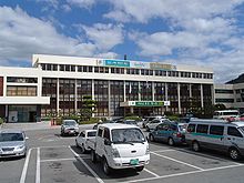 En face d'un bâtiment bas et d'ivoire, un parking et des voitures sont présentés.
