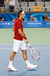 Un homme aux cheveux noirs est dans une chemise rouge avec un short et des chaussures blanches et bandana, où il mène sa raquette de tennis dans sa main droite pointant vers le sol