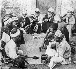 Une vieille photo d'une douzaine d'anciens et hommes d'âge moyen assis sur le sol autour d'un tapis. Un homme en face se trouve à côté d'un mortier et détient une chauve-souris, prêt pour le broyage. Un homme en face de lui détient une longue cuillère.