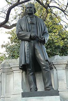 Une sculpture en bronze métallique d'un homme du XIXe siècle portant une longue veste ou un manteau, un pantalon, gilet, avec les outils de dessinateur dans ses mains