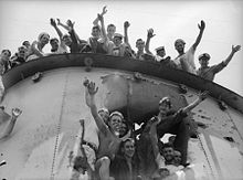 Close up de l'entonnoir d'un navire, qui a un grand trou dans le côté. Les marins sont souriant et saluant le photographe du haut de l'entonnoir et l'intérieur du trou.