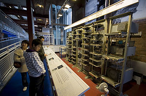 Une série de sept racks métalliques remplis de grands équipements électroniques debout devant un mur de briques. Signes ci-dessus chaque rack décrivent les fonctions exercées par l'électronique qu'ils contiennent. Trois visiteurs des stands d'information lues à partir de la gauche de l'image.