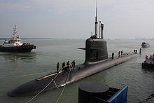 Un sous-marin de classe Scorpène à quai, la moitié hors de l'eau. Les gens sur le dessus sont amarrés, et un bateau peut être vu dans l'arrière-plan