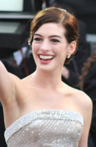 Un large sourire jeune femme, vêtu d'un blanc boiteux robe bustier et de petites boucles d'oreilles, des vagues vraisemblablement à une foule. Son est raie sur le côté gauche et tiré vers le haut et à l'arrière, au large de ses épaules nues. L'arrière-plan est flou et imprécis.
