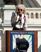 Une femme blonde parlant au kiosque. Elle porte une chemise blanche et lunettes noires. Derrière elle, le balcon d'un bâtiment est visible.