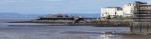 Estructura compatible con patas de metal que salen del mar, que se extiende desde la tierra que muestra varios edificios blancos a una isla con edificios en él. Una casa bote salvavidas con slipway está unido a un lado. En la distancia a través del agua son colinas y edificios.